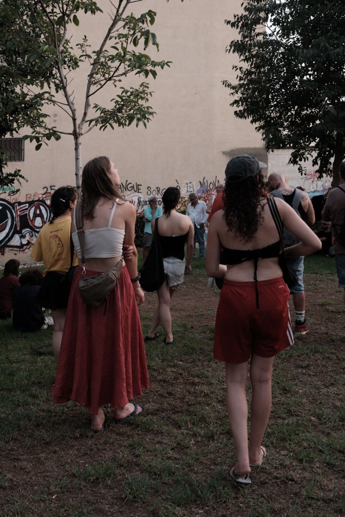 Two women seen from behind walking past a graffiti wall toward a gathered group.