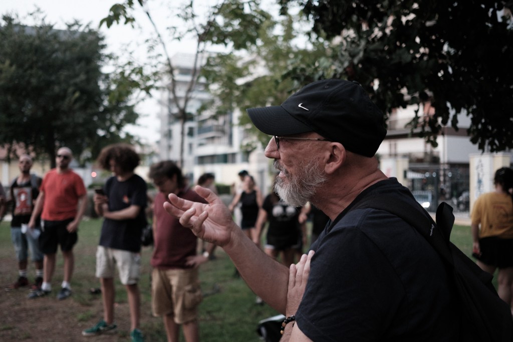 A man in a cap gesturing mid-speech, an out-of-focus street and buildings behind him.