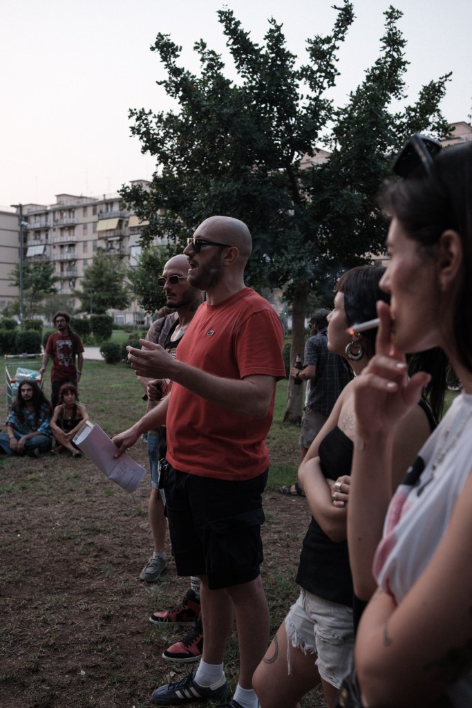 A man in a red shirt speaking with an open hand, others listening and smoking beside him.