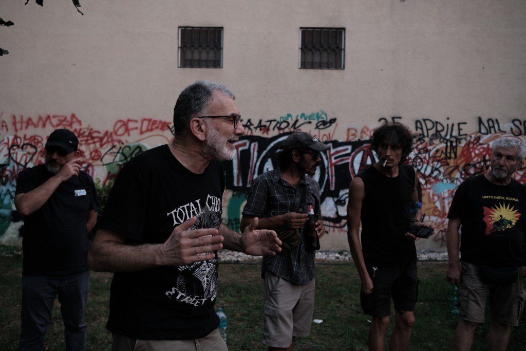 A man mid-sentence in front of a densely tagged wall, others listening around him.