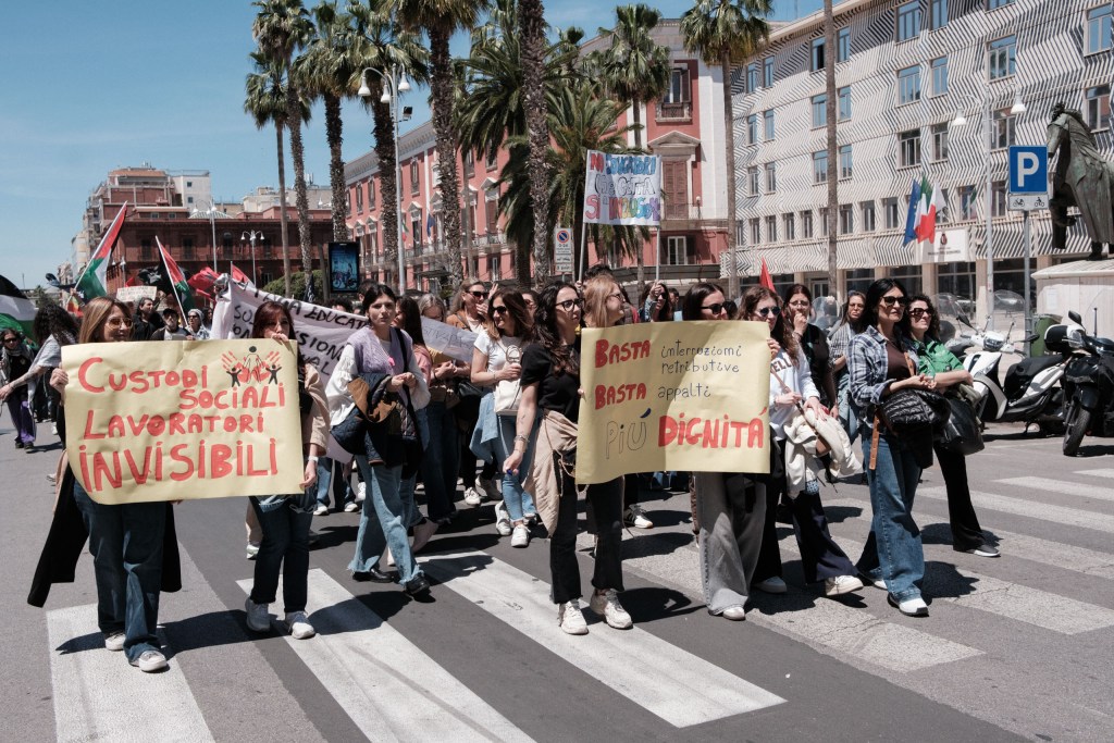 A group of social workers cross a pedestrian crossing carrying signs about outsourcing and workplace dignity.