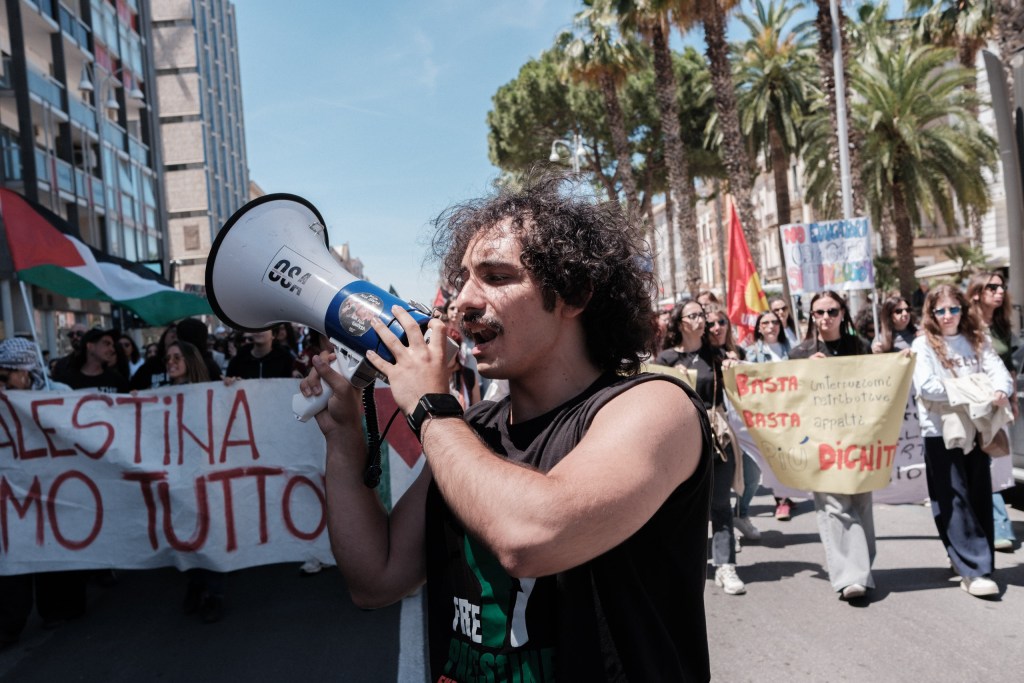 A marcher shouts into a megaphone in front of banners for Palestine and workplace dignity.