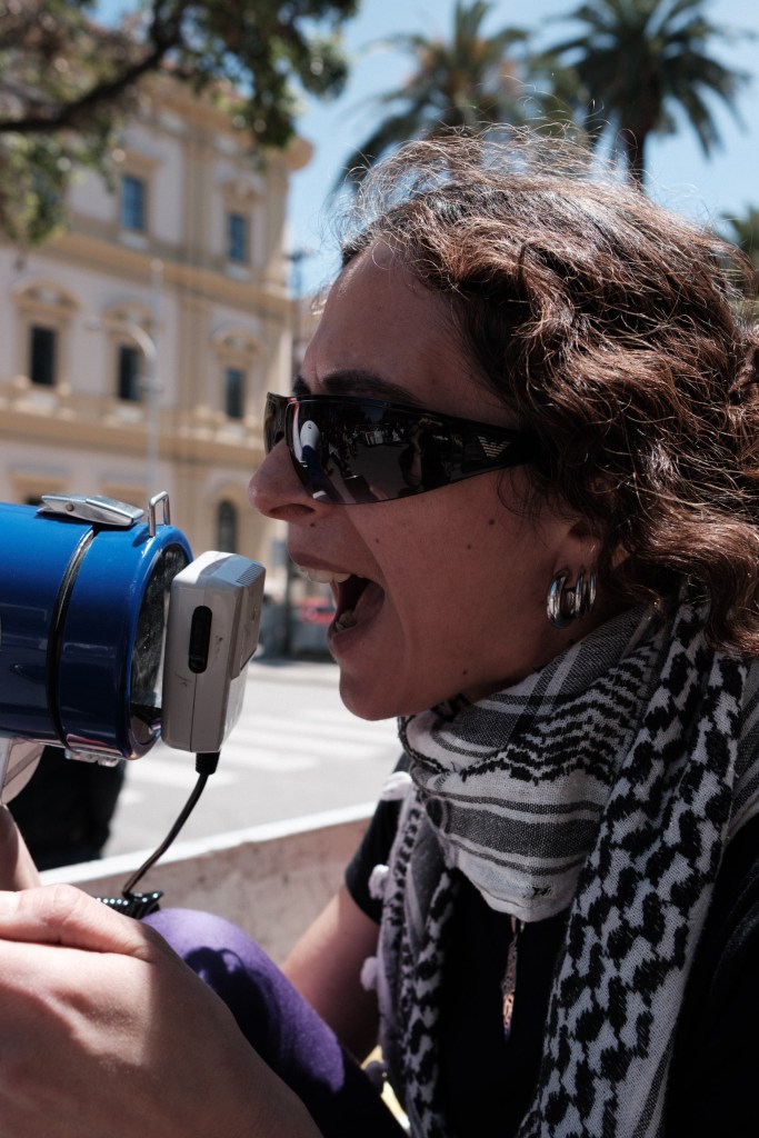Close-up of a marcher with a megaphone, a keffiyeh around her neck, a historic building behind her.