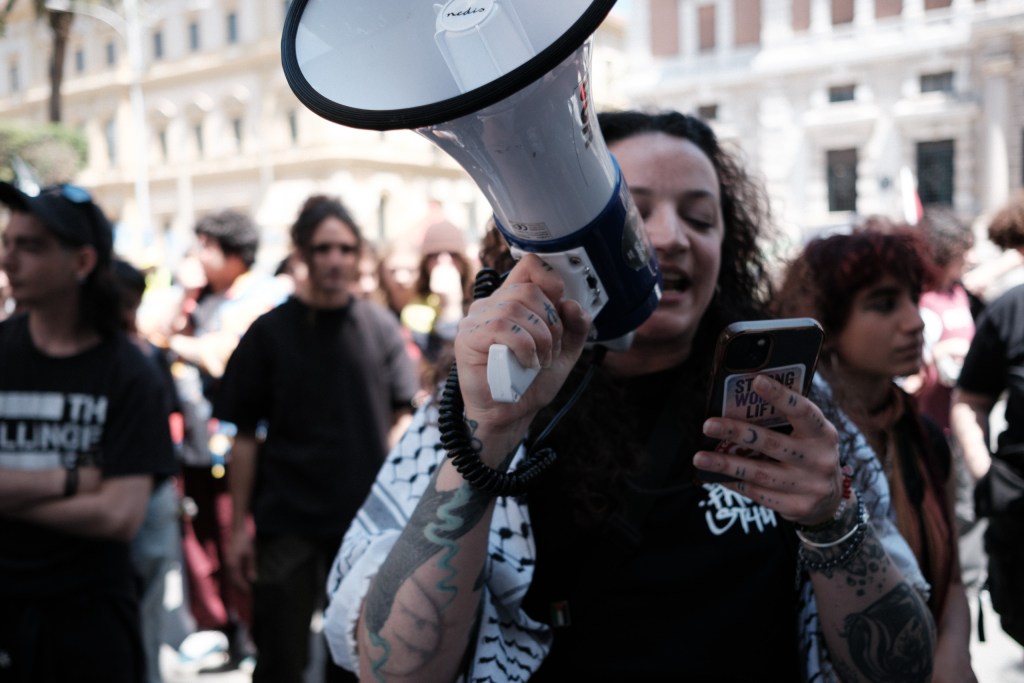 A marcher holding a megaphone and her phone, tattooed forearm, amid the crowd.