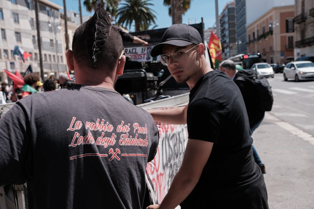 Two marchers exchange a glance before the march sets off, an "Israel is terrorist" banner behind them.