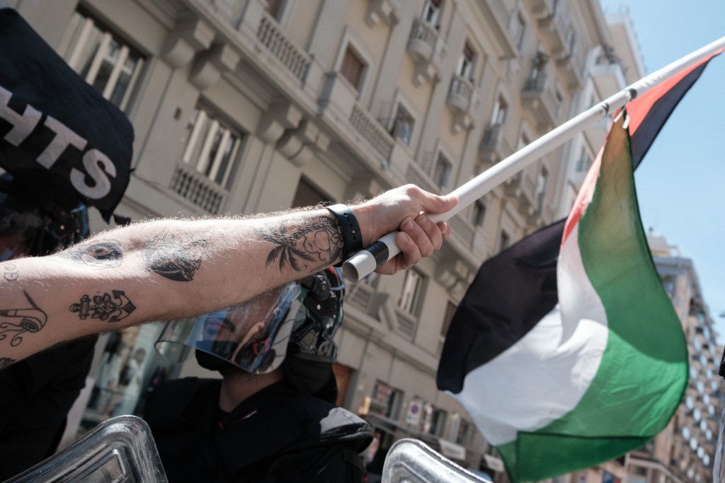 A tattooed arm holds a Palestinian flagpole above a Carabinieri riot shield.