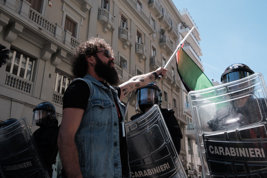 A bearded marcher raises a Palestinian flag in front of the Carabinieri cordon.