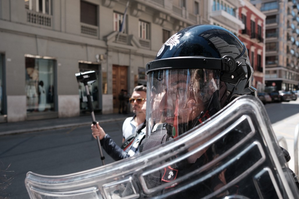 A riot officer watches the scene, a bystander filming with a stabilizer behind him.