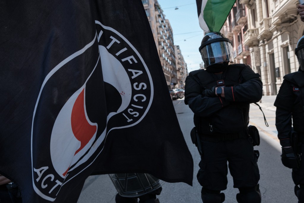 An antifascist flag in the foreground, a Carabinieri officer standing with arms crossed behind it.
