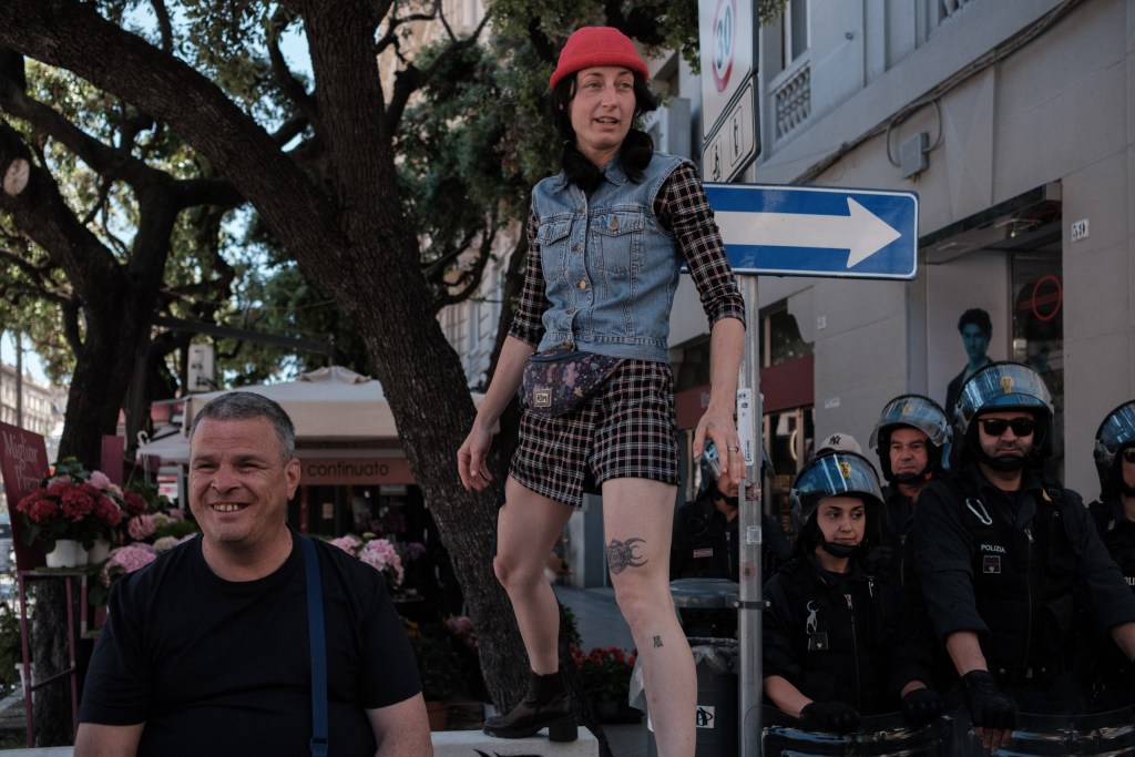 A marcher balances on a bollard, smiling, police officers standing beside her, Via Calefati.