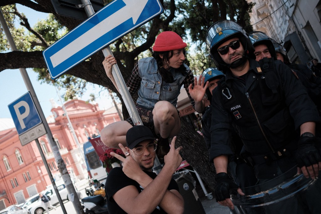 A marcher balances on a bollard, smiling, police officers standing beside her, Via Calefati.