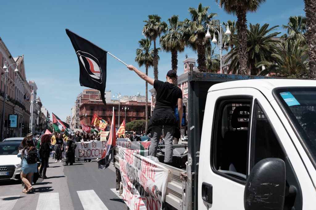 A marcher stands on a van's flatbed raising a flag, the march and palm trees behind him, Bari.