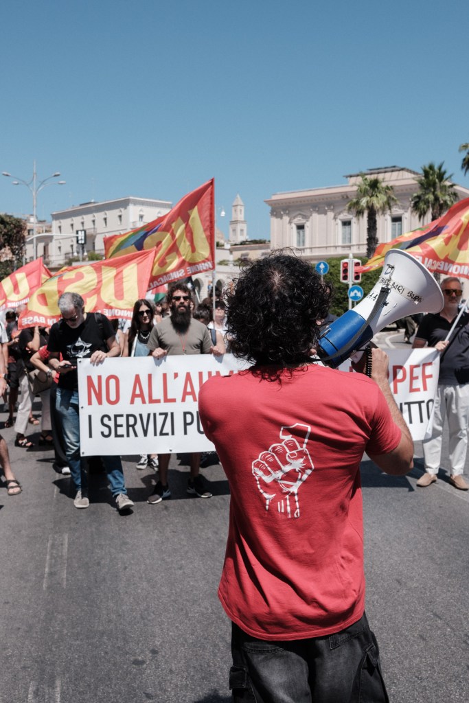 Marcher with a raised-fist t-shirt uses a megaphone at the USB anti-IRPEF rally, Bari, July 10, 2026.