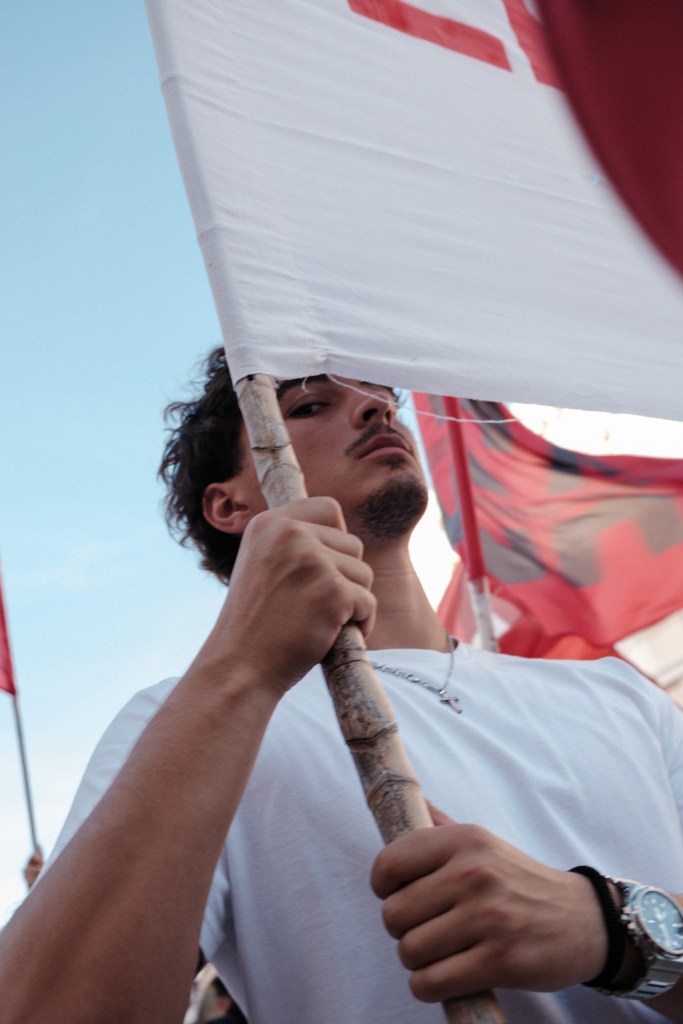 Low-angle view of a young man gripping the cane pole of a white banner, red flags against the sky.