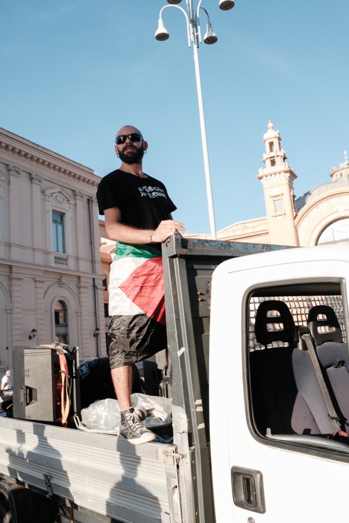 Man with a Palestinian flag tied at his waist stands on the sound truck, the towers of Teatro Margherita in Bari behind him.
