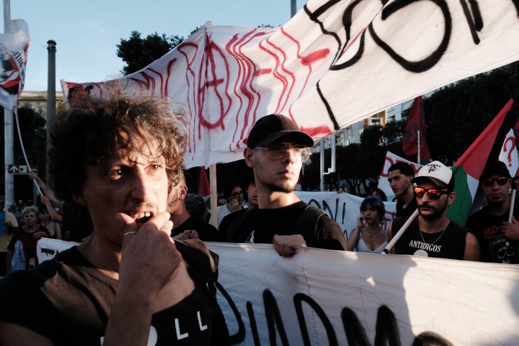 Man blowing a whistle at the head of the march, young protesters holding banners behind him under red spray-painted slogans and Palestinian flags.