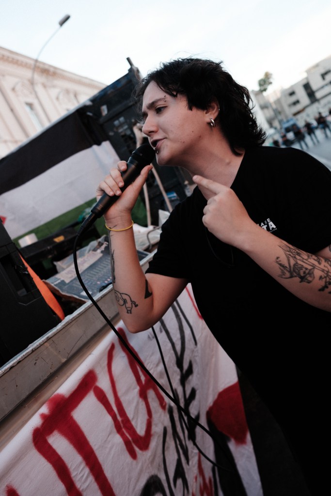 Speaker on the sound truck addresses the crowd by microphone, police vehicles parked down the street.