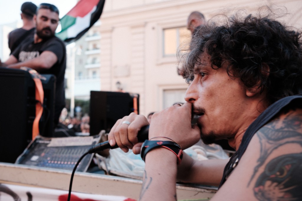 Man pauses with a microphone at his lips on the sound truck, a Palestinian flag behind him.