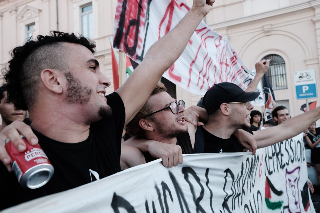 Young protesters arm in arm at the front banner, shouting with fists raised.