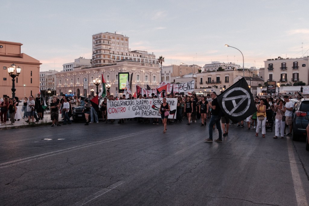The counter-demonstration against the Meloni government advances along a wide Bari road at dusk.