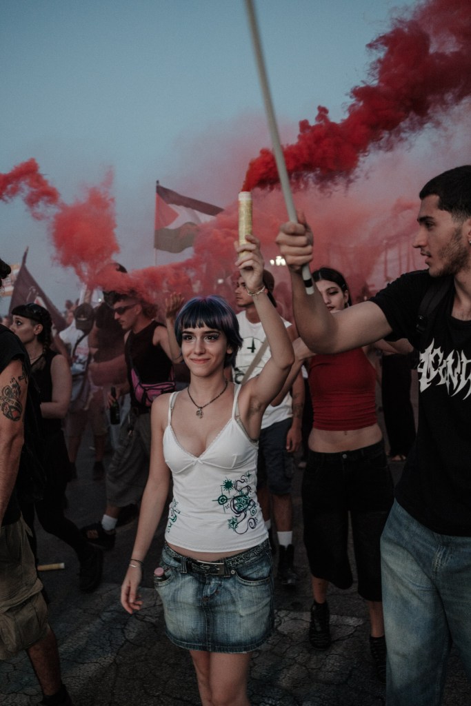 Young woman smiles holding a red smoke flare above her head, a Palestinian flag in the haze behind.