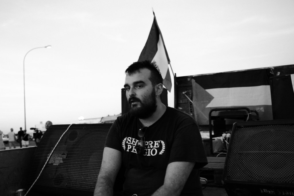 Bearded man seated on the sound truck in a "Sempre peggio" T-shirt, flags hanging over the speakers.