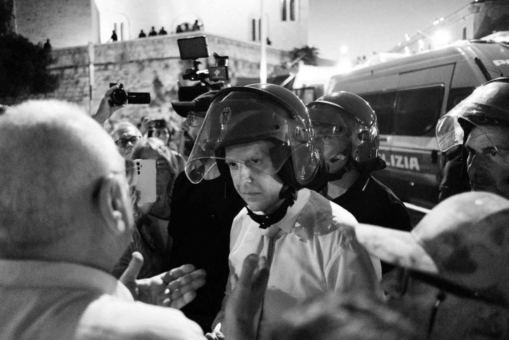 Officer in a white shirt and riot helmet faces protesters at close range while phones record.