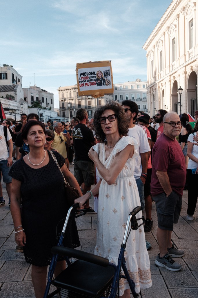 Two women in the crowd, one leaning on a rolling walker, below a hand-drawn sign mocking Meloni over diesel prices.