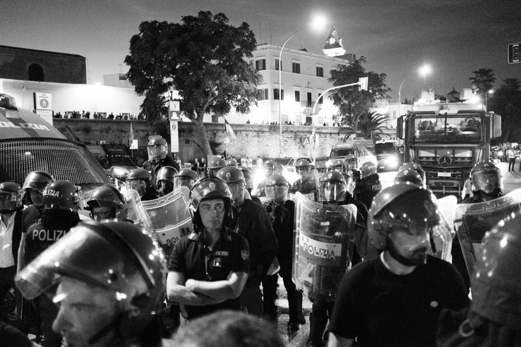 Riot police line with shields blocks the Bari march at night, people watching from the old walls above.