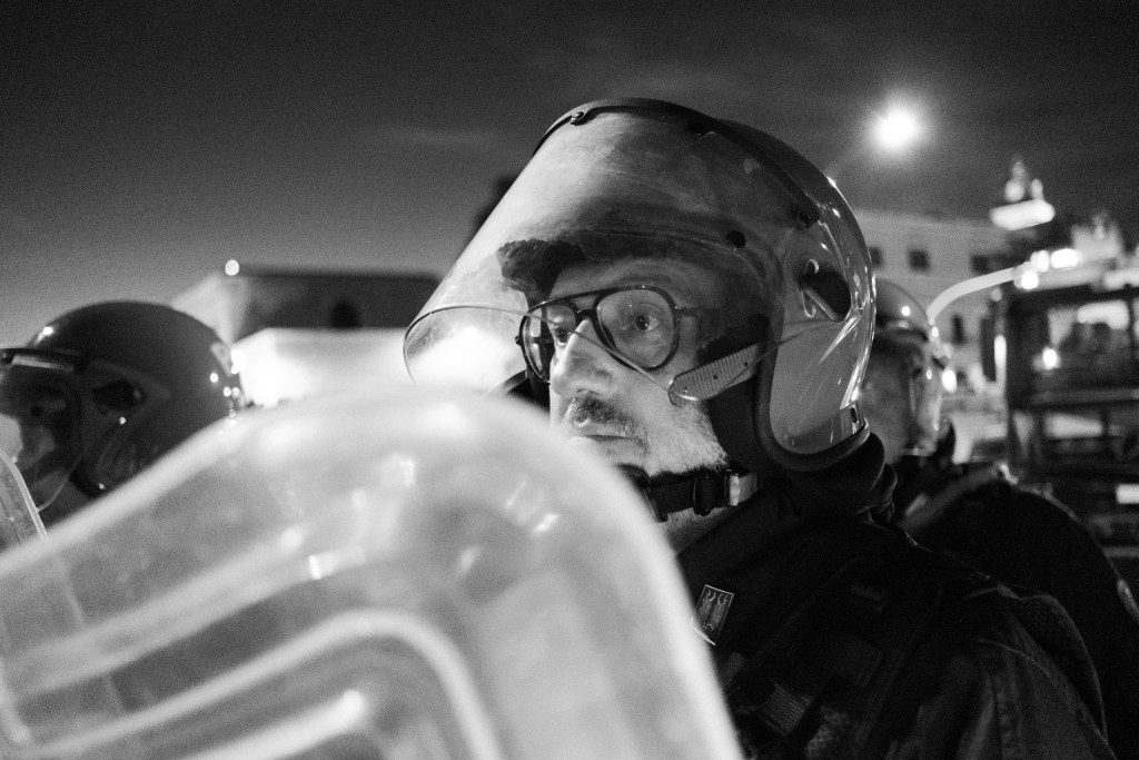 Officer in glasses behind a raised visor, eyes on the crowd.