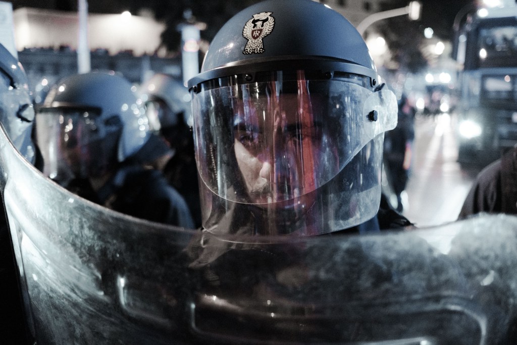 Officer behind a riot visor and shield, street lights reflected in the plastic.