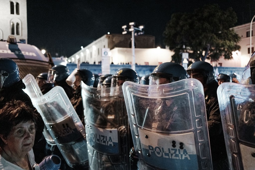 Older woman stands in front of a wall of police shields, a lit stone bell tower behind the line.