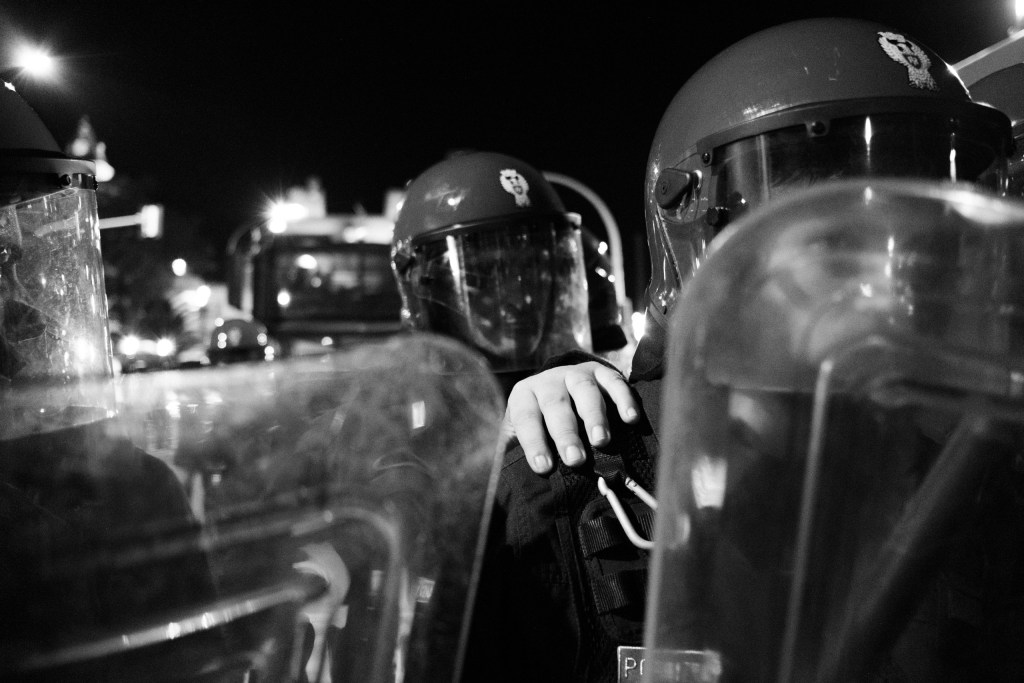 One officer's hand resting on another's shoulder between riot shields.