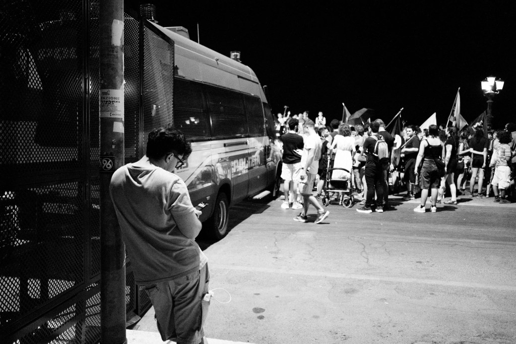 Young man leaning on a pole checks his phone beside a police van, the crowd a few steps away.