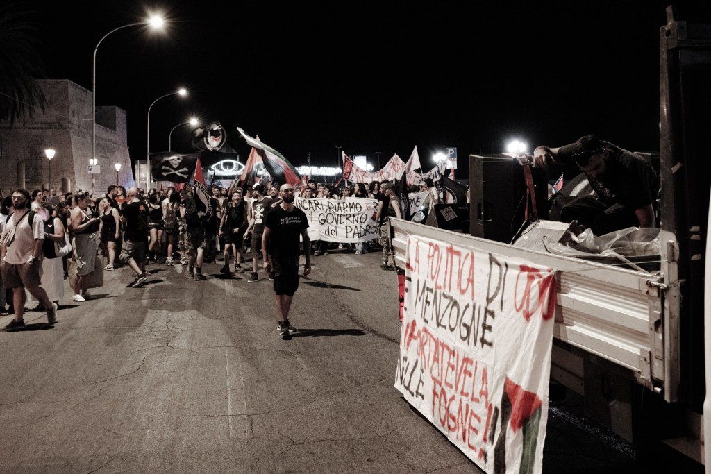 The march leaves the seafront at night past the stone fortification, a "politica di odio e menzogne" banner on the sound truck.
