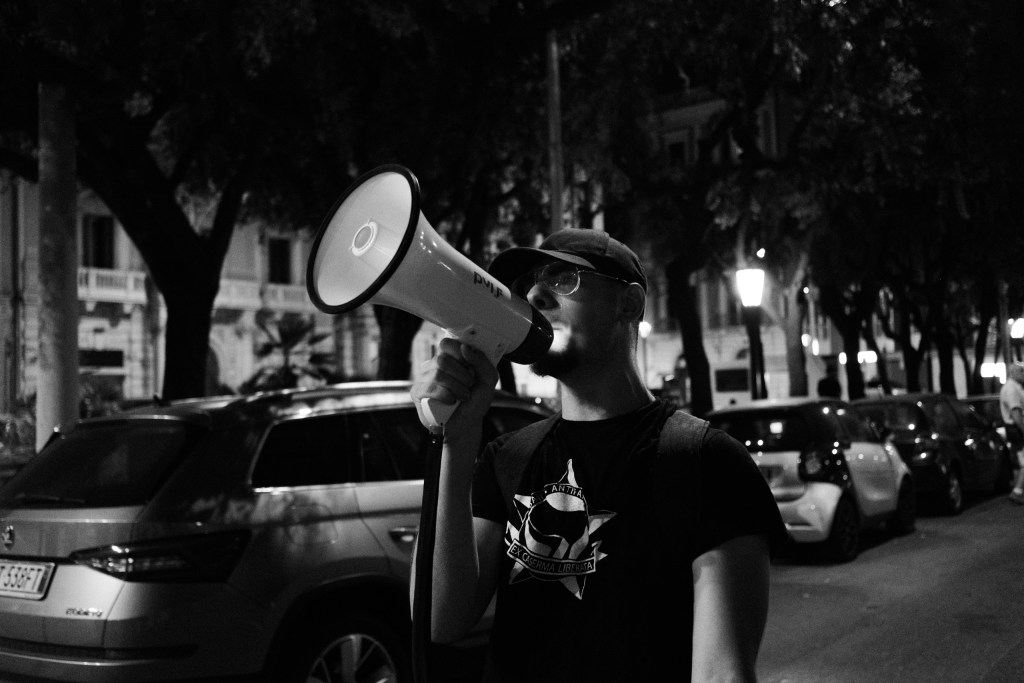 Protester in an Ex Caserma Liberata T-shirt shouts through a megaphone on a tree-lined street at night.