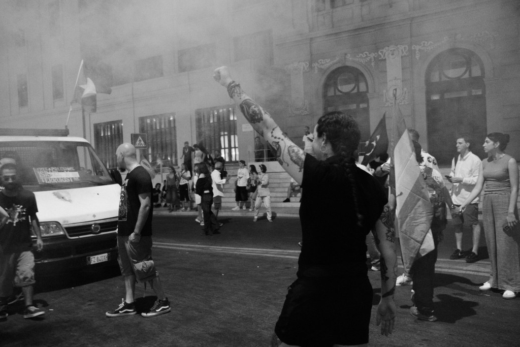 Woman with a tattooed arm raises her fist through smoke in the street in front of Teatro Margherita.