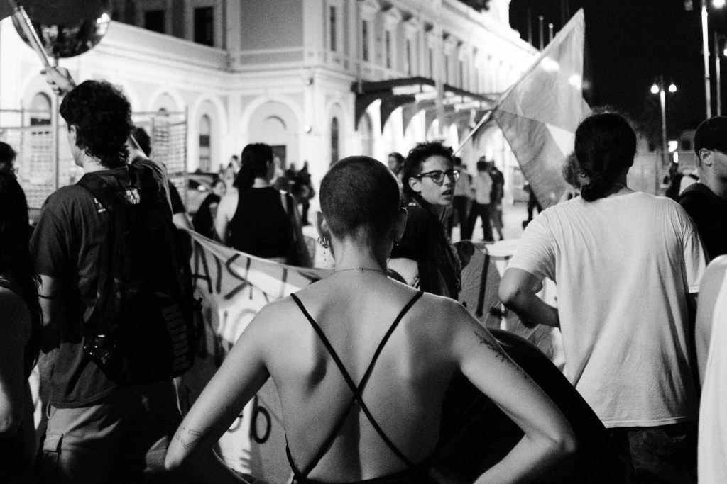 Protester seen from behind, shaved head and crossed straps, as the march reaches Bari Centrale.