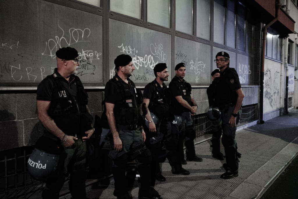 Officers in berets wait against a graffiti-covered wall, helmets hanging at their sides.