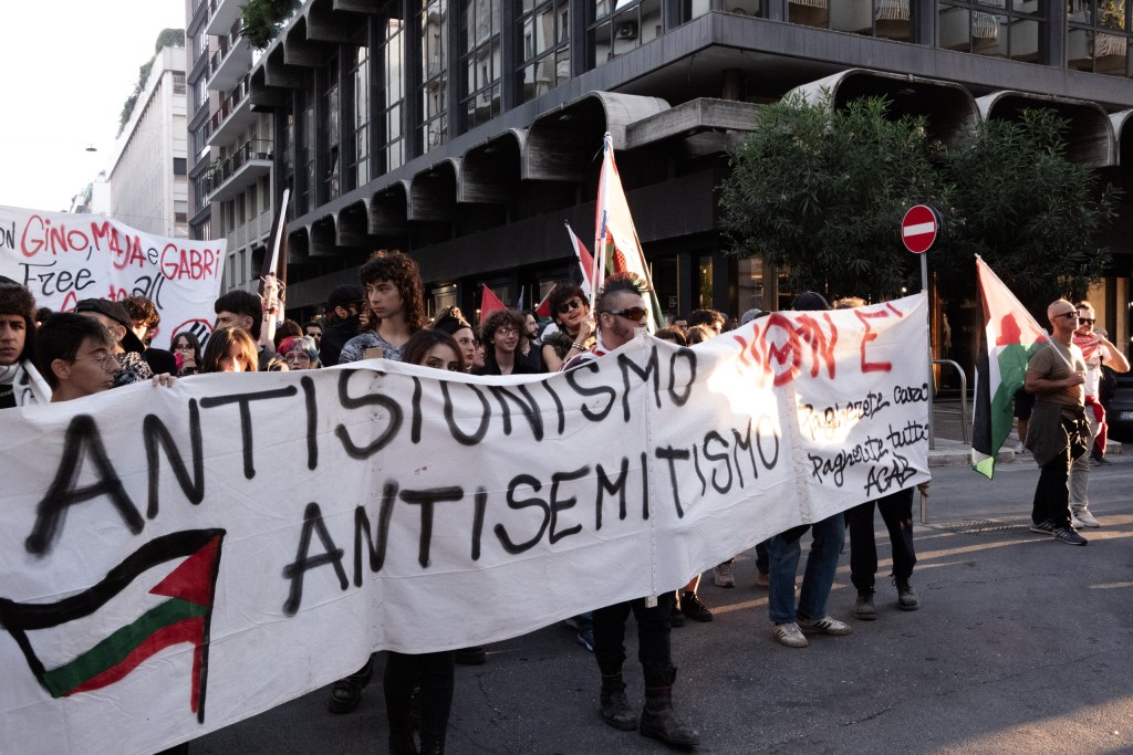 Demonstrators march along a city street in low late-afternoon light, holding a large white banner that reads "Antisionismo non è antisemitismo"; Palestinian and red flags rise behind them.