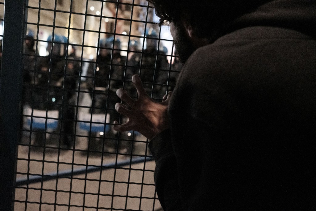 A man seen from the side, his hand pressed against a metal mesh barrier, faces a line of riot police in blue helmets lit by street lights.
