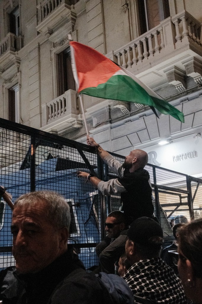 A man raised above the crowd waves a Palestinian flag over a metal barrier, with a blue police vehicle behind it; demonstrators in the foreground look on.