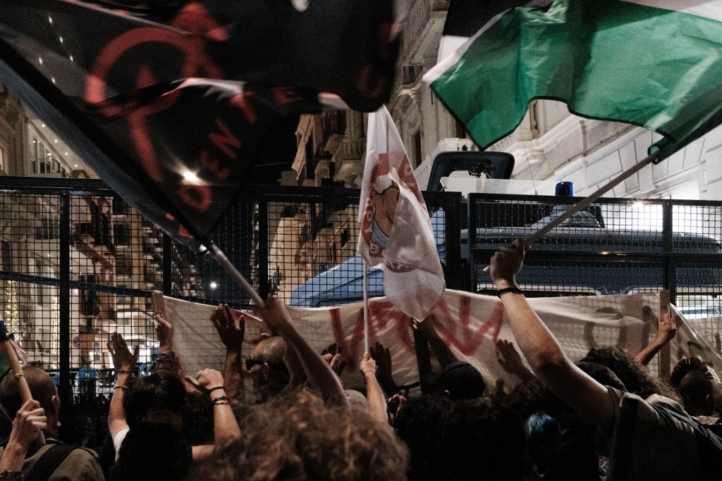 Raised hands reach toward a banner held against a barrier, under black, white and green flags; a police van with a blue light stands behind the fence.