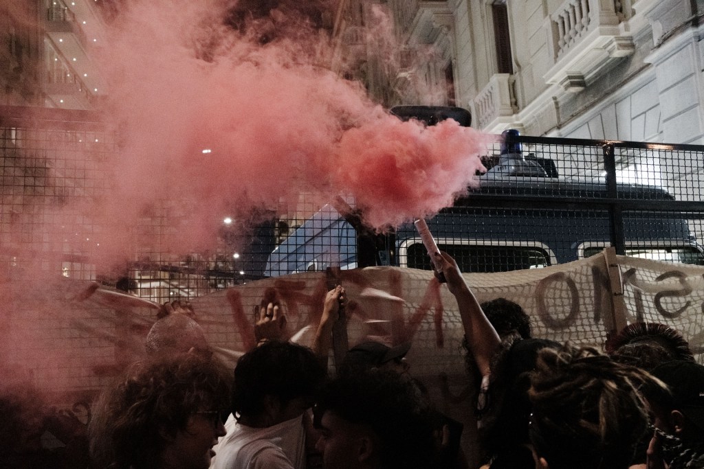 A red smoke flare is held up above a banner at the barrier; smoke fills the left of the frame, and a police van with a blue light is visible behind the fence.