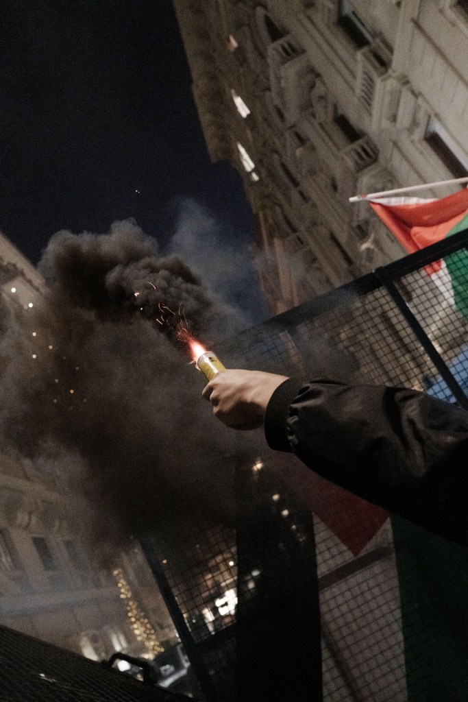 A hand holds a lit flare that throws sparks and black smoke upward between buildings, next to a metal barrier and a Palestinian flag.