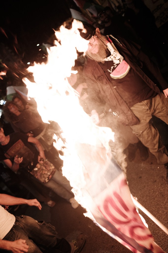 An Israeli flag on a pole burns in the street; a bearded man in a keffiyeh stands behind it and other demonstrators watch.