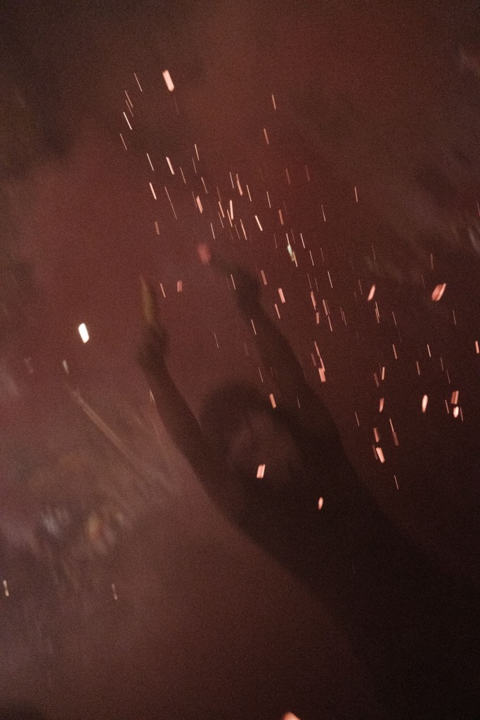 Sparks streak upward through a dark red haze; the blurred silhouette of a raised arm is visible behind them.