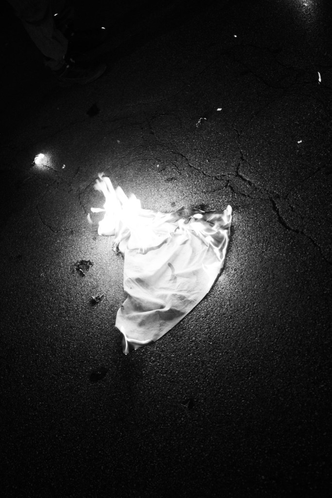 A piece of Israeli flag lies on dark asphalt, lit by its own flames.