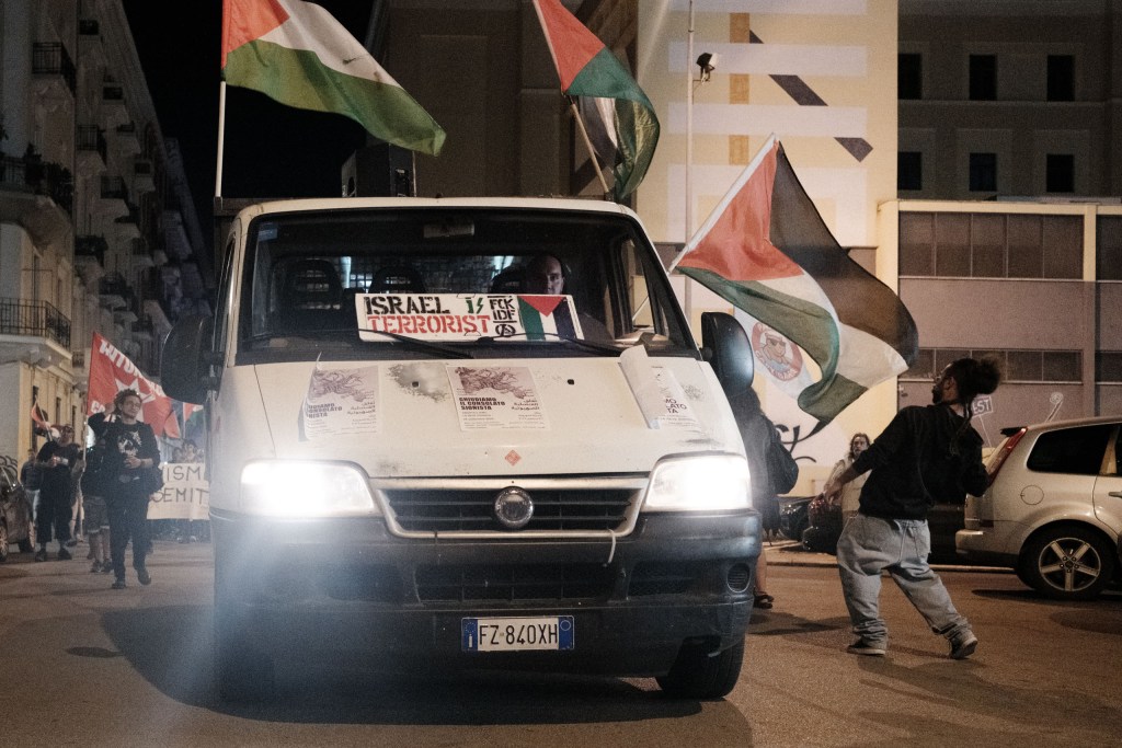 At night, a white truck with its headlights on moves along the street, marchers visible behind it; a sign on the windscreen reads "Israel terrorist", flyers on the hood read "Chiudiamo il consolato sionista" in Italian and Arabic, and Palestinian flags fly above.