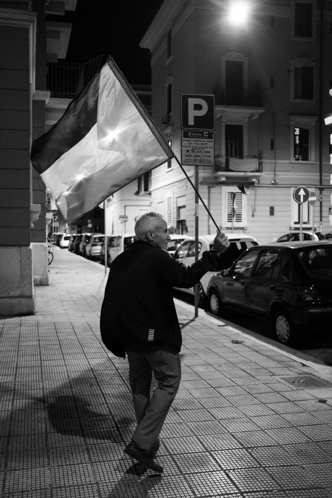 An older man with white hair walks along a night-time sidewalk waving a Palestinian flag, past parked cars and a parking sign.